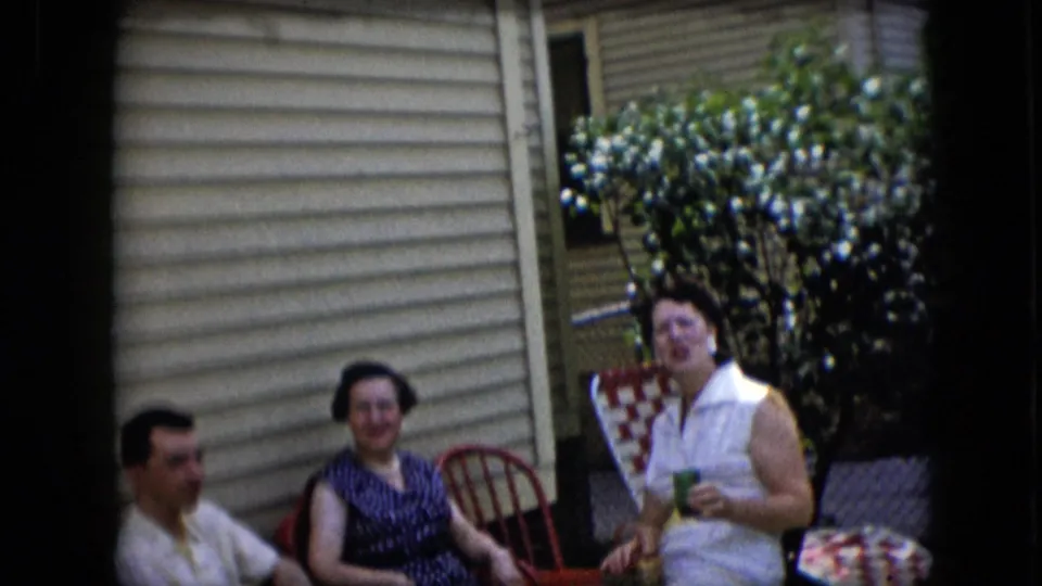 California 1969 home-movie frame showing adults seated outside a home, with a woman in a white sleeveless dress smiling beside red and checkered patio chairs.
