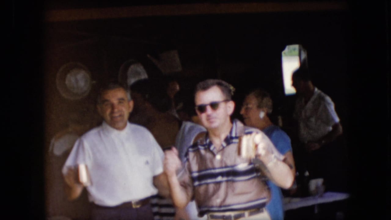 1969 California 8mm frame showing adult men and other guests gathered near a shaded garage area, with one man in a striped polo and sunglasses holding a drink.