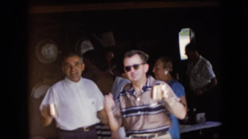 1969 California 8mm frame showing adult men and other guests gathered near a shaded garage area, with one man in a striped polo and sunglasses holding a drink.