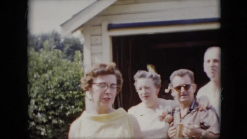 1969 California 8mm frame showing adults standing in bright daylight outside a garage, including women in light summer clothing and a man wearing sunglasses.