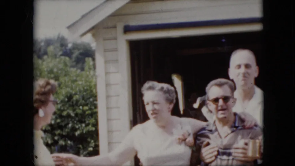 1969 California 8mm frame showing adults gathered near an open garage doorway, with a woman in a light dress beside a man in sunglasses holding a drink.