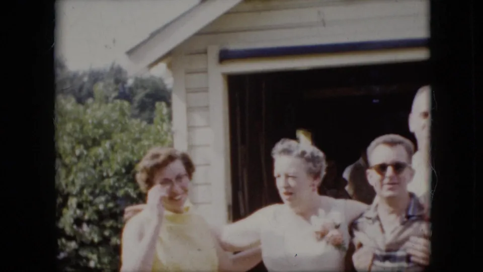 1969 California 8mm frame showing smiling adults standing close together in front of a garage, with a man in sunglasses holding a beverage and women in light summer clothing.