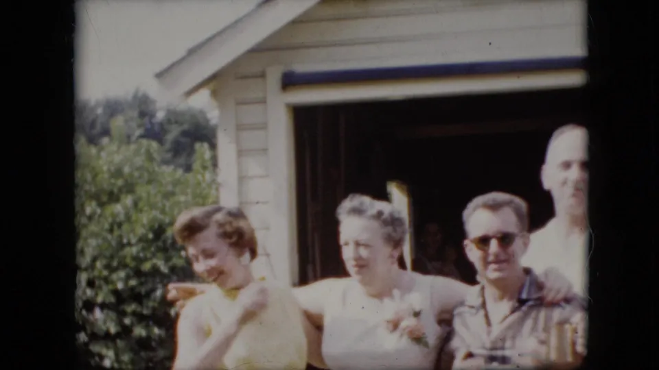 In 1969 California home-movie footage, four adults pose outside a light-colored garage as one woman in a yellow dress looks away and another woman stands beside two men.