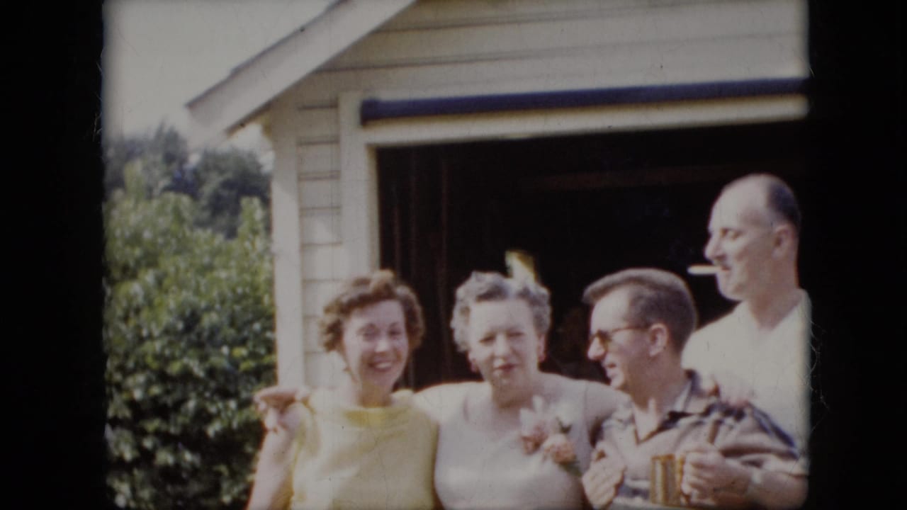 Two women and two men smile and pose near an open garage, with one man holding a cigarette.