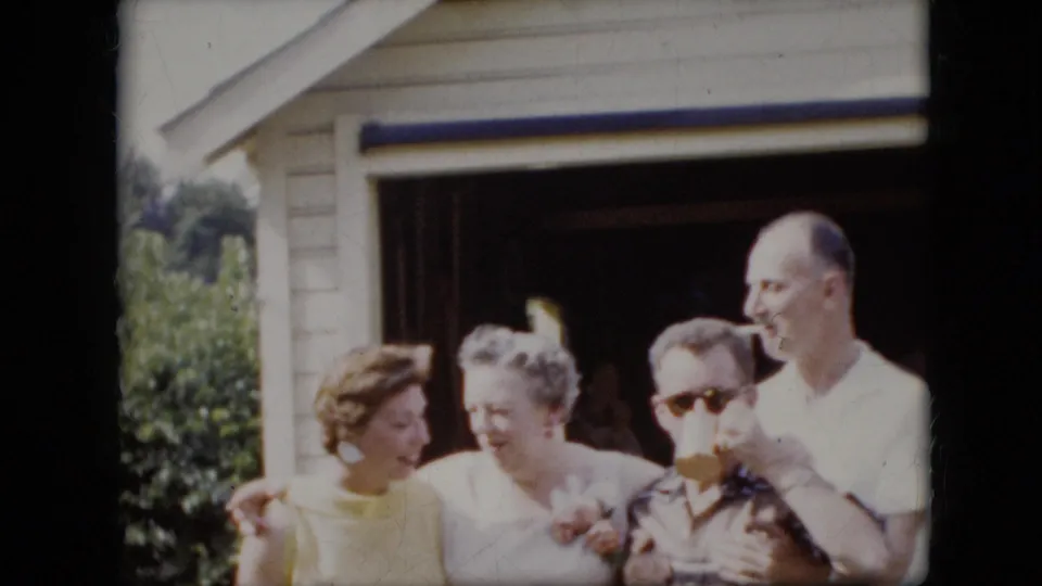 The 1969 California home-movie frame shows adults clustered in bright sunlight outside a garage, including a woman in yellow, a woman in white, a man in sunglasses drinking, and a man smoking.