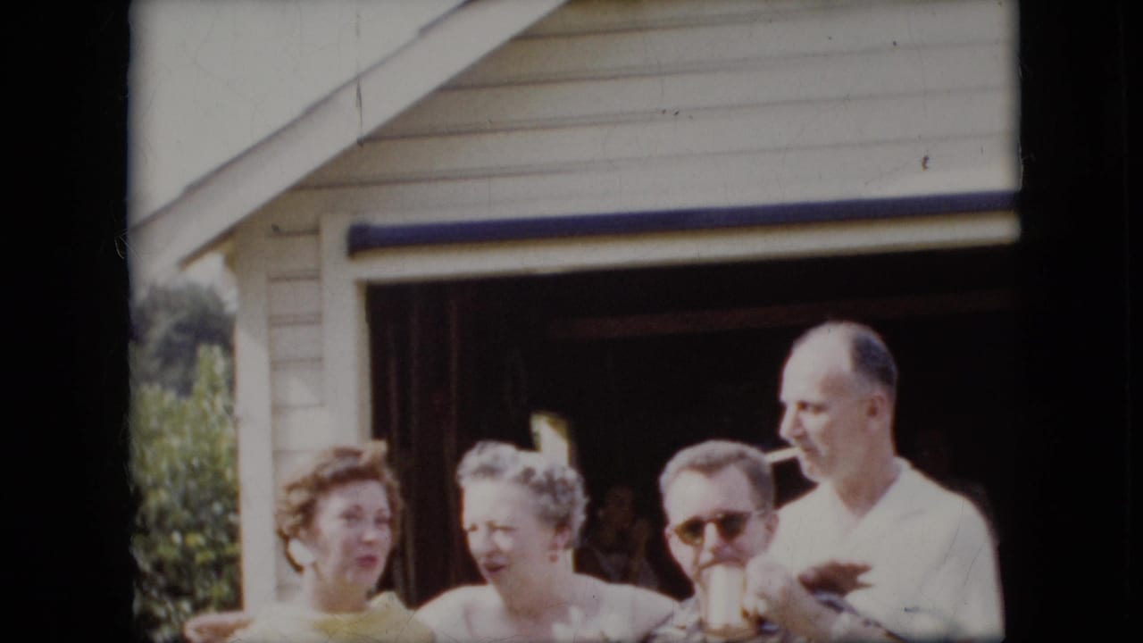 In this 1969 California 8mm frame, two women and two men stand outside a suburban garage with arms interlocked, with one man wearing sunglasses and another holding a cigarette.
