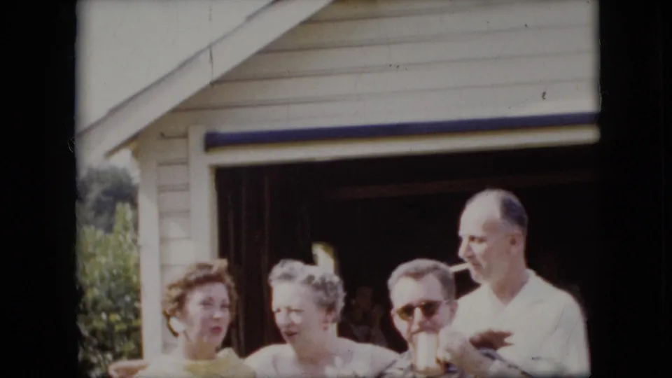 In this 1969 California 8mm frame, two women and two men stand outside a suburban garage with arms interlocked, with one man wearing sunglasses and another holding a cigarette.