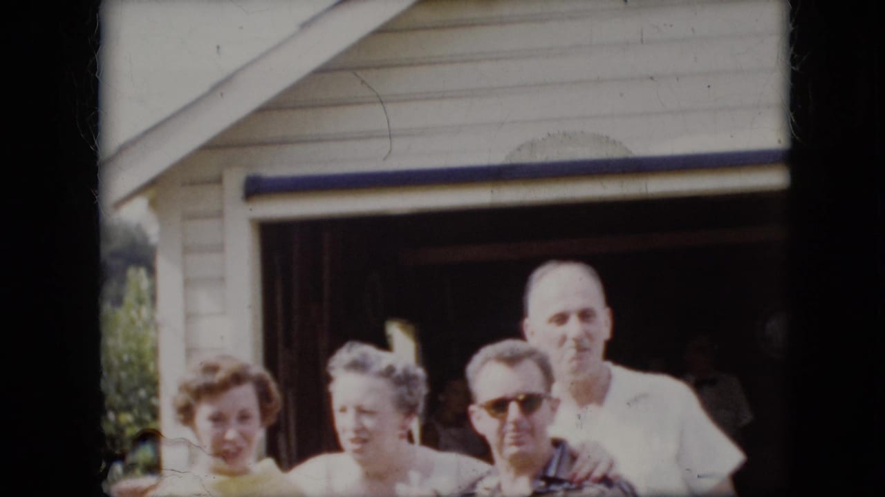 In this 1969 California home-movie frame, four adults face the camera outside an open garage, including a woman in a yellow dress, an older woman in light clothing, a man in sunglasses, and a man in a white shirt.