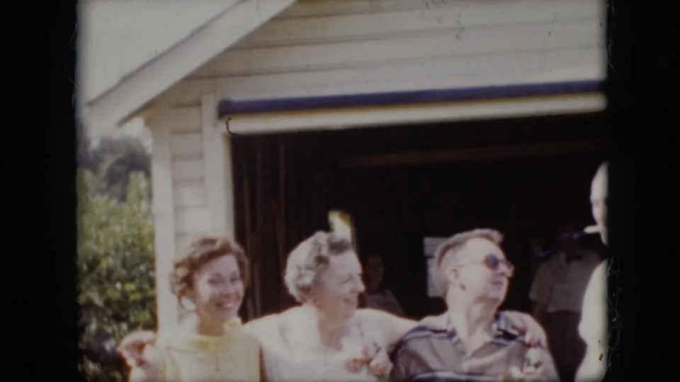 This 1969 California 8mm frame shows adults laughing and leaning together outside a suburban garage, with the man in sunglasses turned toward the group and another figure partly outside the frame.