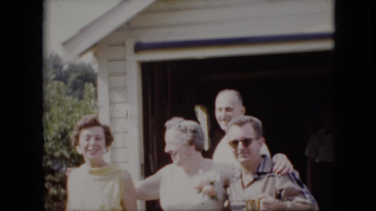 Four adults smile together outside a light-colored house in bright daylight.