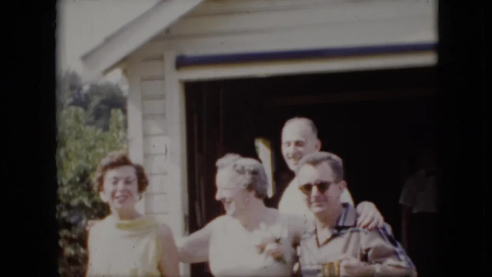 Four adults appear outside a suburban California house in supplied 1969 home-movie footage, with one seated man in sunglasses and others smiling toward the camera.
