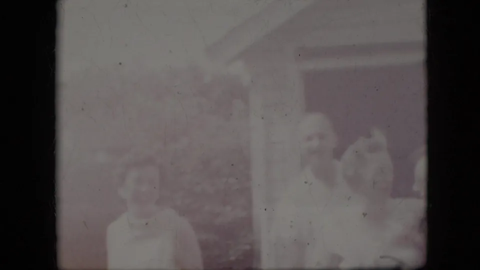 A heavily faded 8mm frame shows adults near the same house exterior, with faces and body outlines visible but softened by exposure and color loss.