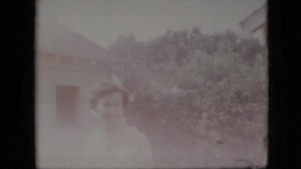 A washed-out home-movie frame shows a partial adult figure in front of shrubs and a house exterior, with details obscured by fading and exposure.