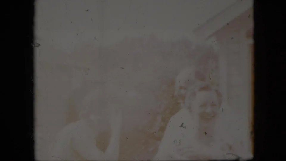Faded 1969 California home-movie frame showing faint seated family members outdoors, with a smiling older woman visible near the right edge amid soft focus and film wear.