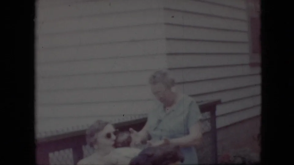 Faded 1969 California home-movie frame showing horizontal siding, a porch rail, an indistinct adult-shaped figure, and a small animal- or child-sized shape near the lower left.