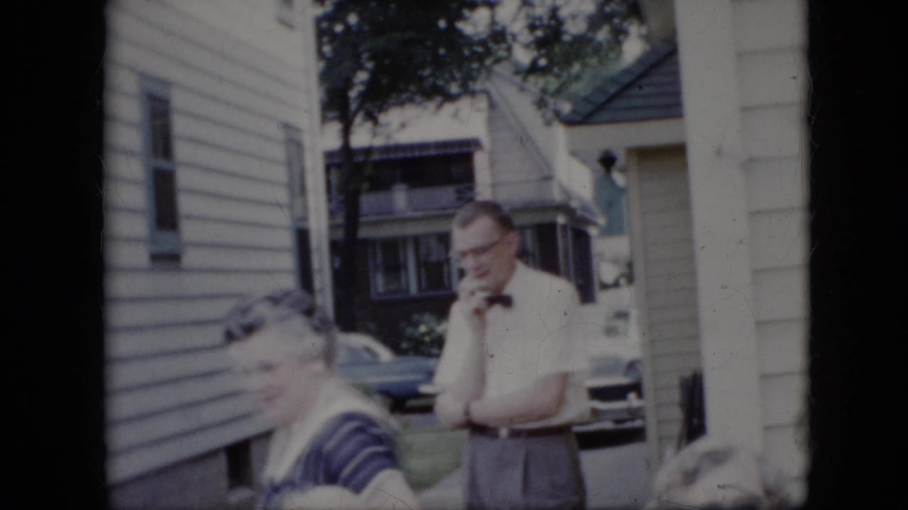 Adults gather outside a suburban house while a man in a bow tie stands near the driveway.