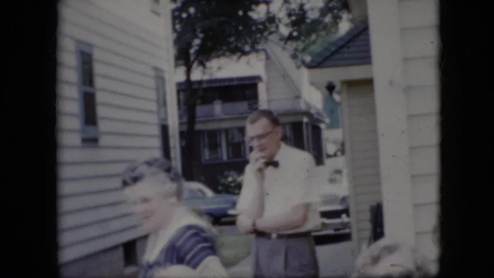 1969 California home-movie frame showing adults outside a light-sided suburban house, with a man in a white shirt and bow tie standing behind seated family members.
