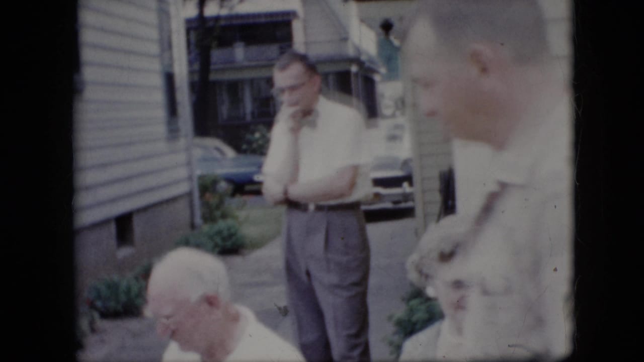 Several adults sit and stand outside a house, including a man in a bow tie with his hand near his face.