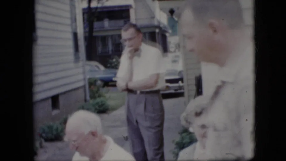 1969 California home-movie frame with a standing man in a bow tie, seated adults in the foreground, house siding, windows, greenery, and parked cars behind them.
