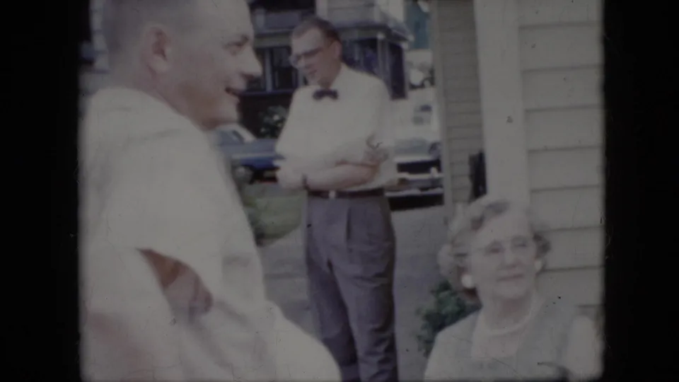 1969 California home-movie frame showing a smiling older man in the foreground, a seated woman, and a man in a bow tie standing near a driveway outside a suburban home.