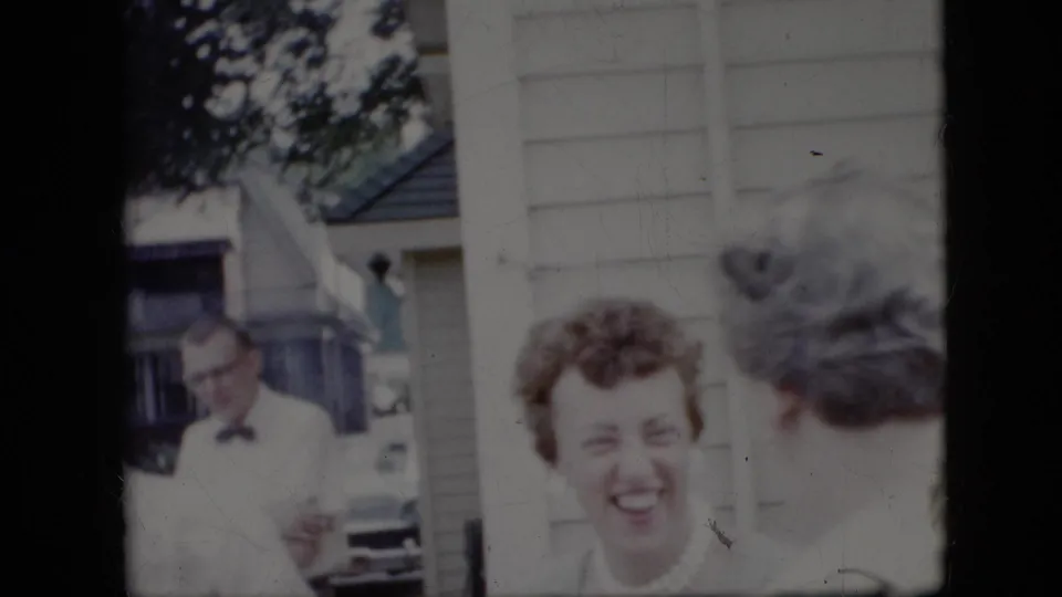 1969 California home-movie frame with a smiling woman in the foreground, another woman seen from behind, and a man in a bow tie partly visible near a suburban house and driveway.