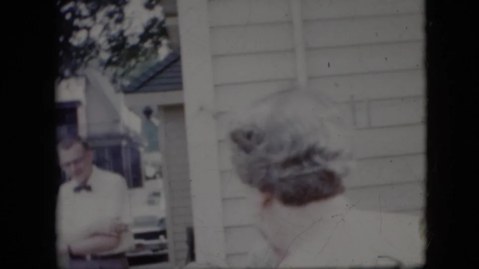 In 1969 California 8mm home-movie footage, a man wearing a bow tie stands with arms crossed near other adults outside a light-sided residential house.