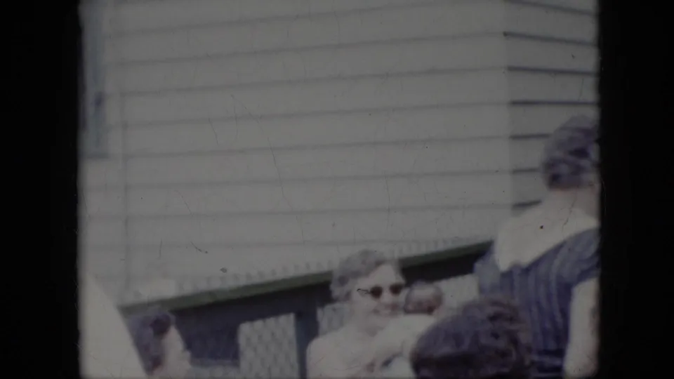 A woman wearing sunglasses appears to smile or laugh beside other adults near a porch or deck railing in the California backyard gathering.