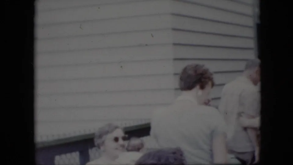 Adults in casual clothing stand close together beside the light-sided house, with a woman in sunglasses partly visible at the lower edge of the frame.