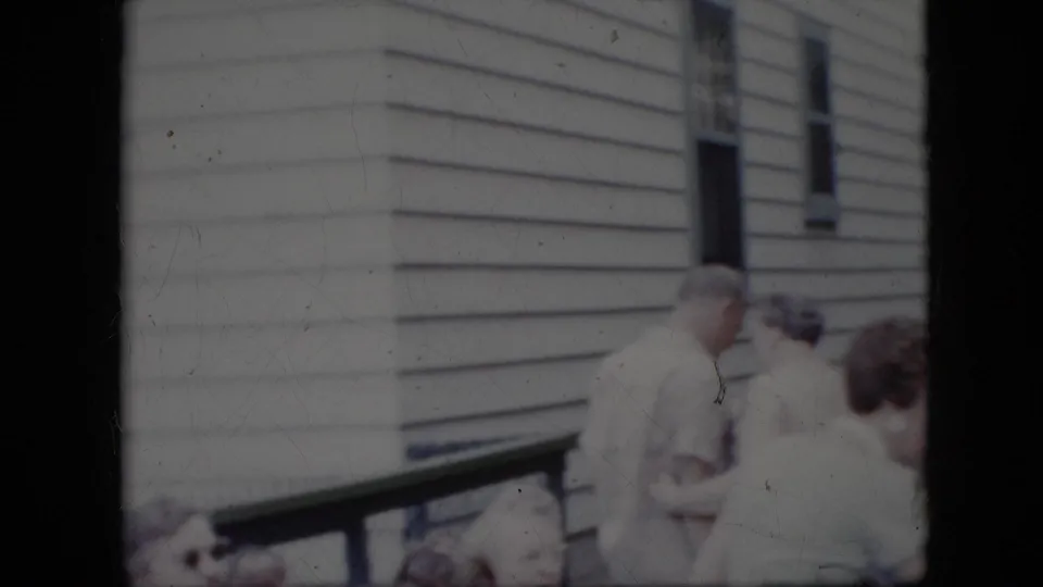 Several adults stand near a wooden railing outside the light-sided house, continuing the informal 1969 California backyard social scene.