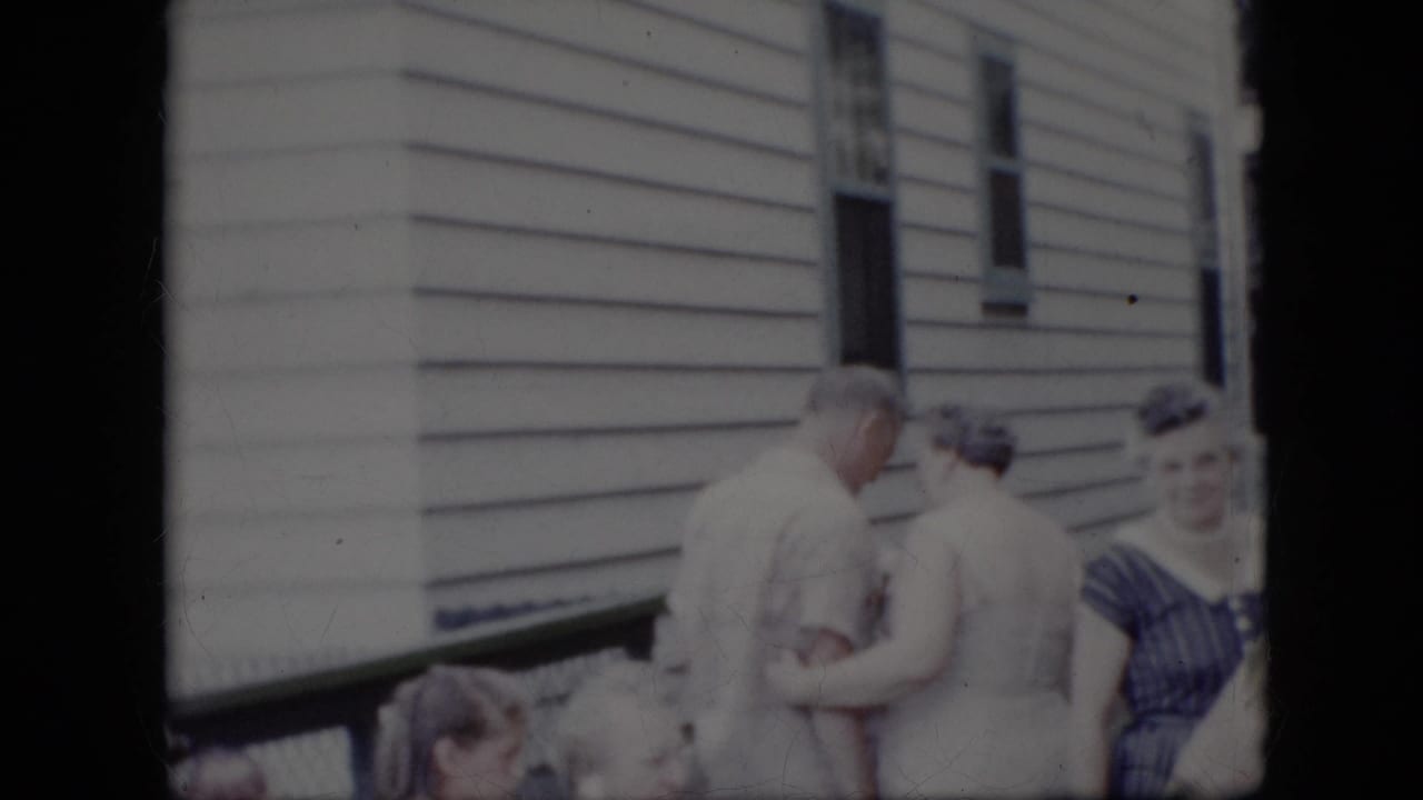 Adults gathered outside a white-sided house, with a man leaning toward a woman near seated people.