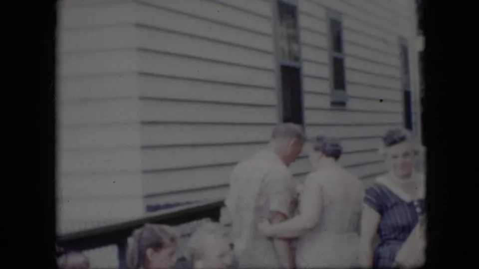 In metadata-supported 1969 California home-movie footage, adults gather outside a white-sided house as a man in a light shirt leans toward a woman while others stand and sit nearby.