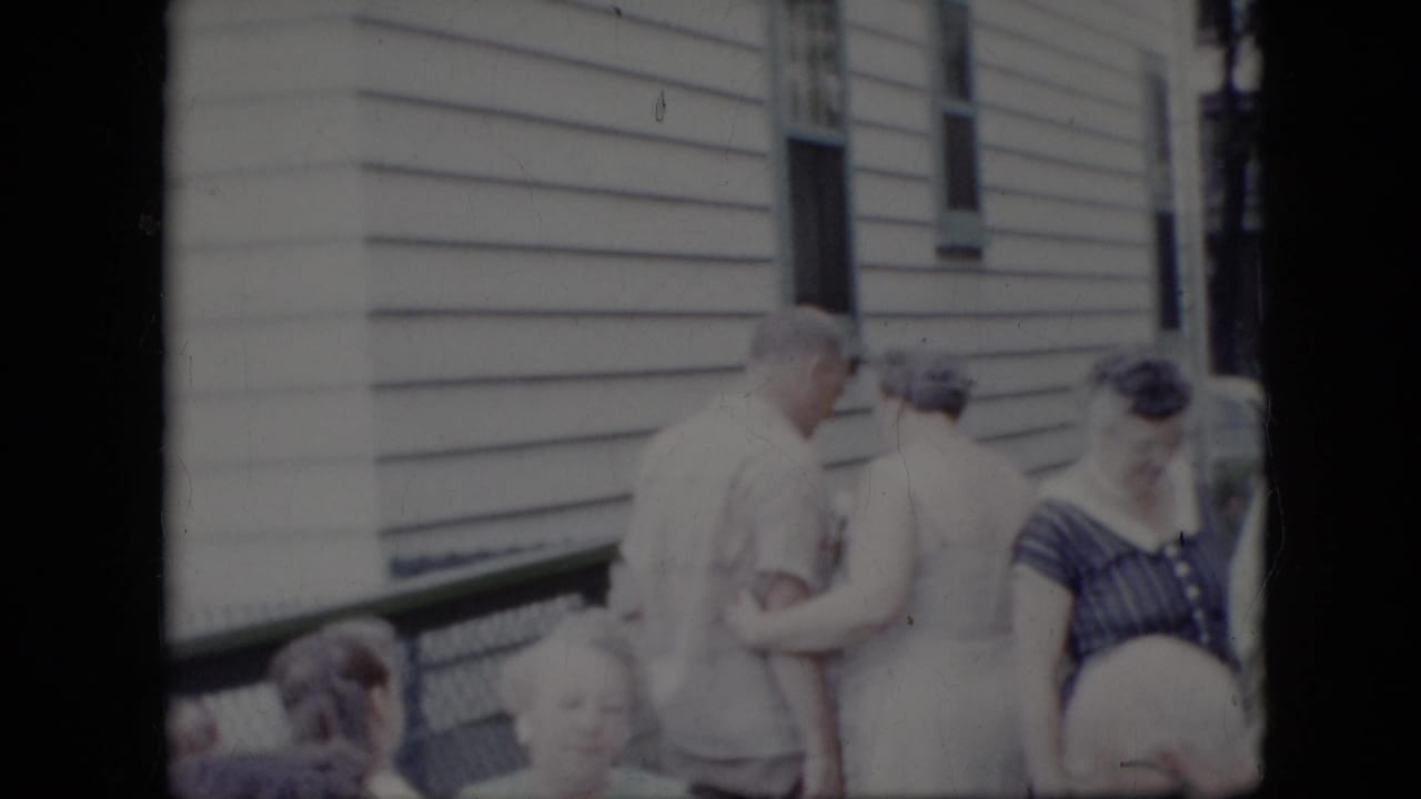 People stand and sit outside a white-sided house while a man and woman greet each other.