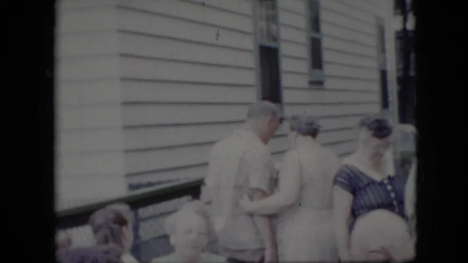Adults in late-1960s attire stand and sit outside a white-sided house in California, with a man and woman close together in a casual greeting.