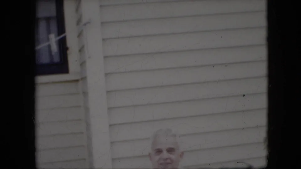 The home-movie frame shifts lower on the white-sided house, showing an exterior window and a partial close view of an older man's face near the bottom edge.