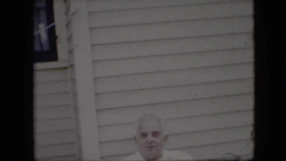 An older man is partly framed beneath the white siding of a house in this metadata-supported 1969 California home-movie shot.