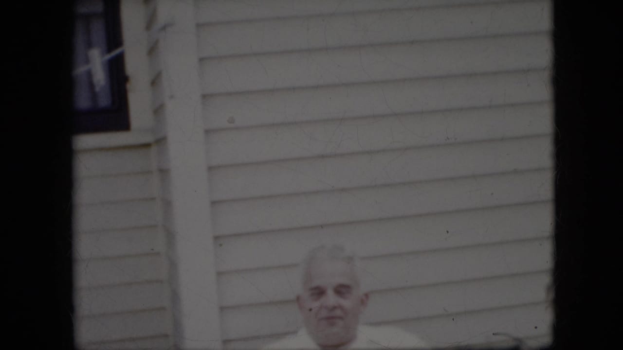 Older white-haired man in a white shirt seated before a light-sided house.