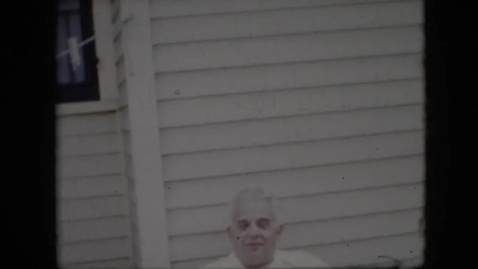 An older white-haired man in a white shirt sits outdoors in front of a light-colored house with horizontal siding, in 1969 California home-movie footage.