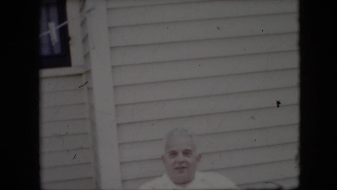Older man facing the camera with house siding and a window behind him.