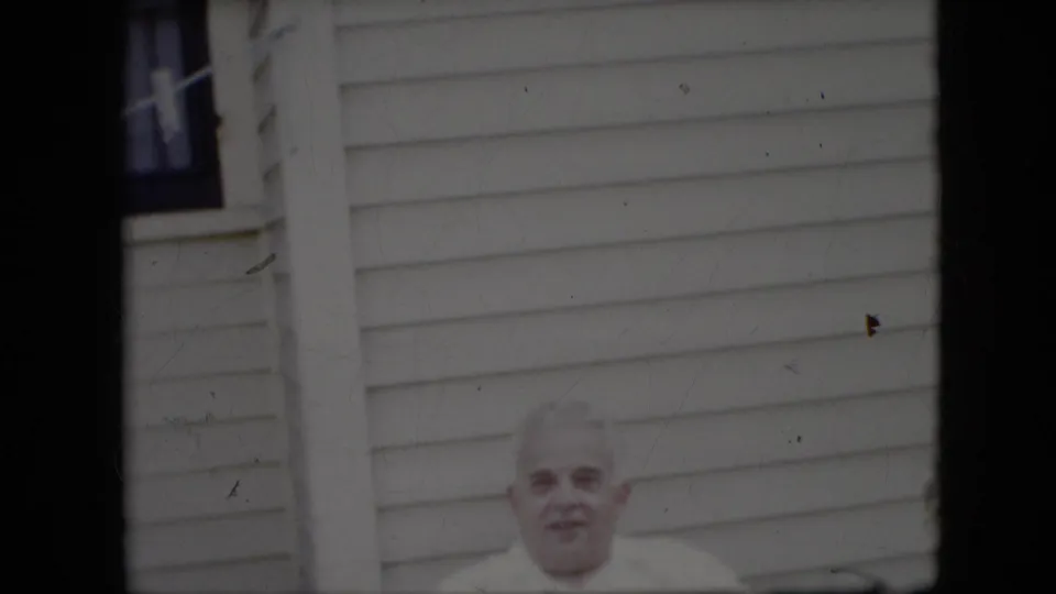 The older man faces the camera from a seated position, framed low against pale horizontal siding and a narrow window in the 1969 California home-movie scene.