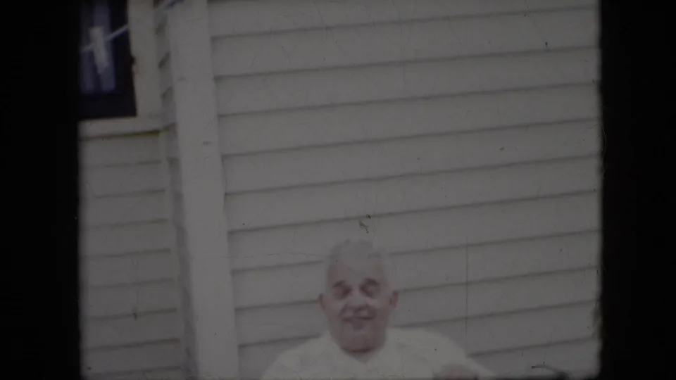 The older man appears lower in the frame, seated beneath light horizontal siding with dark film borders and soft archival color fading.