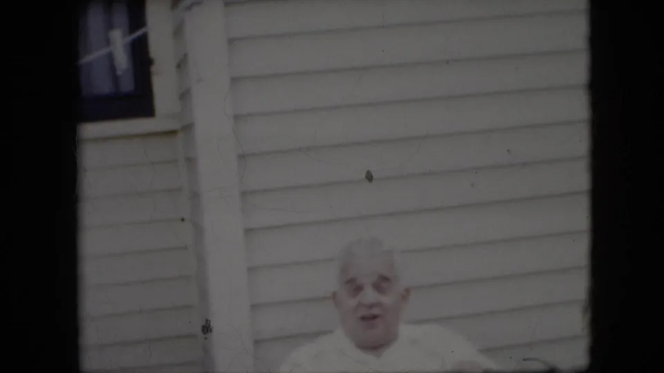 An older white-haired man in a white shirt remains seated outside, partially low in the frame against pale house siding in the California 1969 home-movie clip.