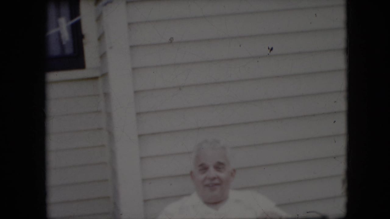 Elderly man in a white shirt smiling outside a light-sided house.