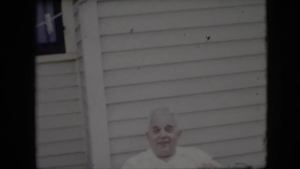 In metadata-supported 1969 California home-movie footage, an elderly man in a white shirt smiles toward the camera in front of light horizontal house siding.