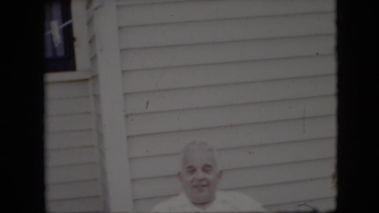 Elderly man seated outside with light siding behind him.