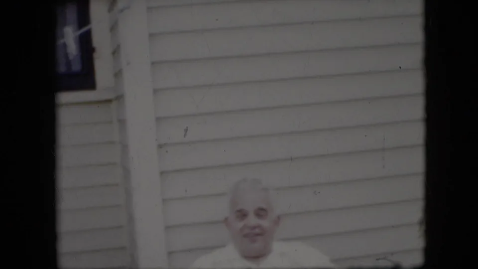 The elderly man remains framed low in the shot, wearing a white shirt and looking toward the camera against the exterior siding of a house.