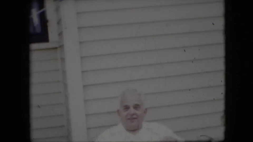 A soft Super 8-style frame shows the elderly man lower in the composition, facing the camera with light-colored exterior siding filling most of the background.