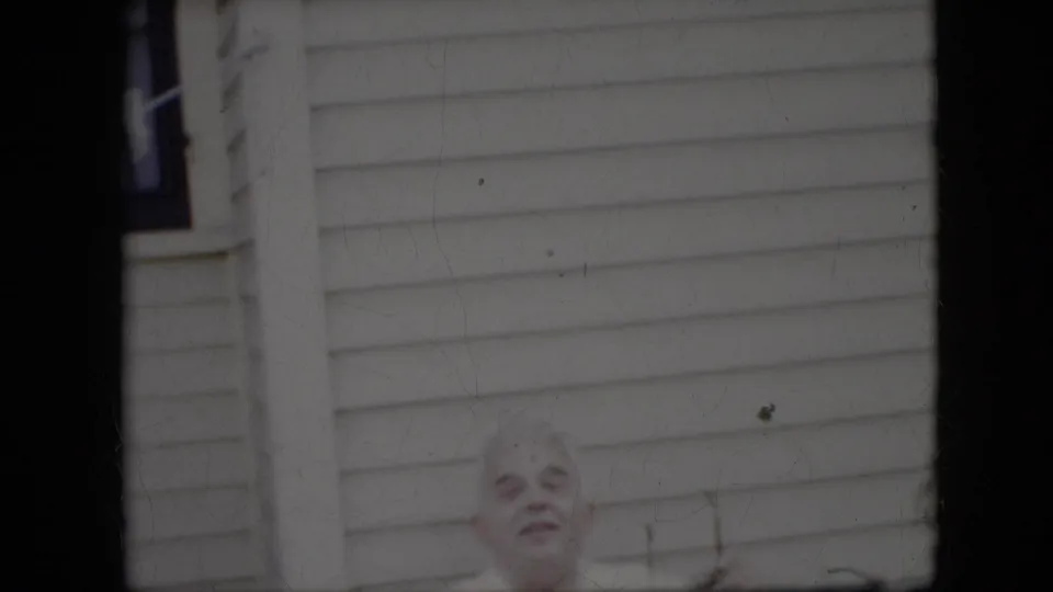 In this California 1969 home-movie frame, a white-haired elderly man appears near the lower edge in front of a light-sided residential exterior.