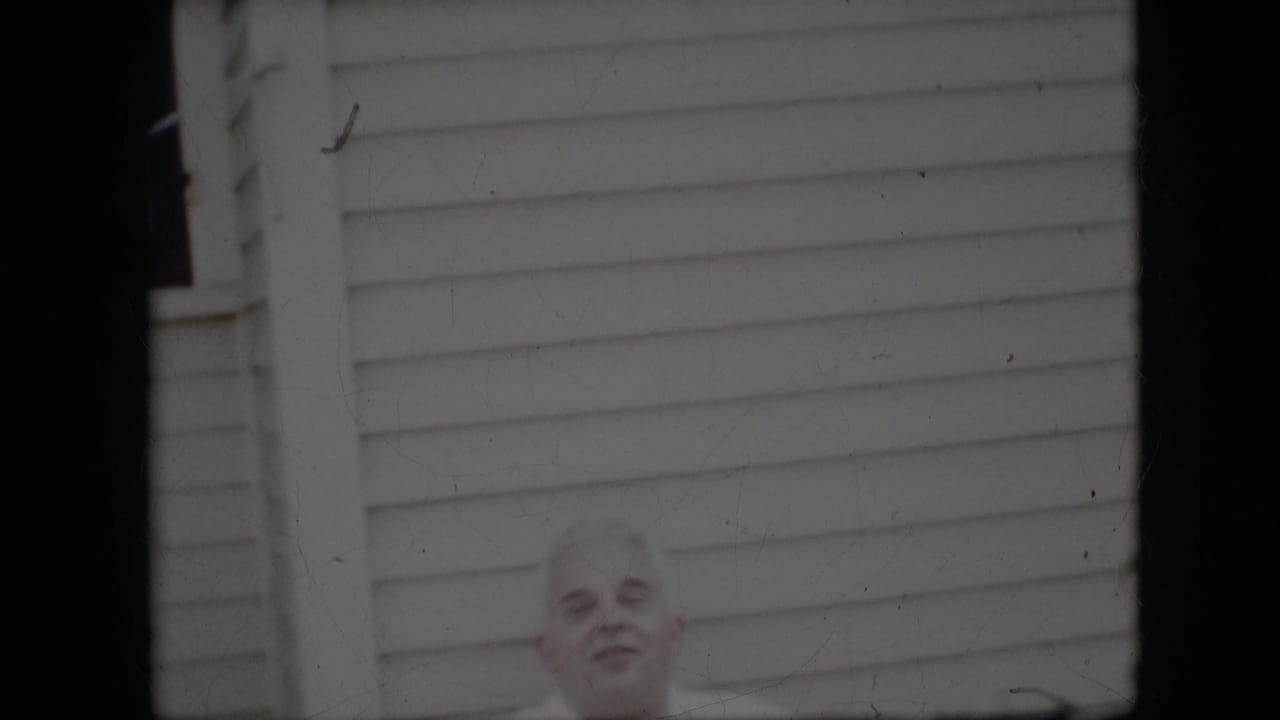 Elderly man standing low in frame against white exterior siding.