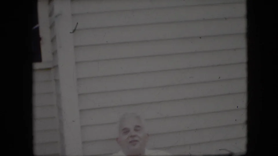 The elderly man remains partly framed against white house siding, with soft focus, dark film edges, and visible dust from the 8mm scan.