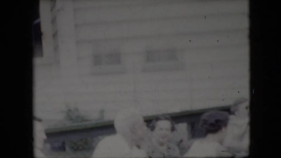 Adults gather outdoors below the light-sided house, with the older man and other family members partly visible in a soft, faded 8mm frame.